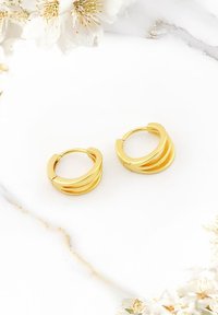 Goldene Creolen mit drei geschichteten Abschnitten, glatter Textur und runder Form. Vor einem hellen Marmorhintergrund mit floralen Akzenten präsentiert.