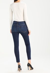 Jeans ajustados de mezclilla en azul oscuro, con una cintura de altura media, diseño de cinco bolsillos y detalles de costura sutiles. Combinados con tacones negros.