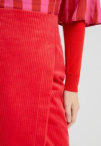 Rode corduroy rok met verticale ribbels, gecombineerd met een effen rode lange mouwen shirt. Close-up van een hand op de rok.