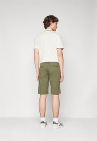 Polo a maniche corte da uomo in bianco sporco con rifiniture scure, abbinato a pantaloni corti verde oliva. I pantaloni corti presentano due tasche posteriori e una sottile grafica.