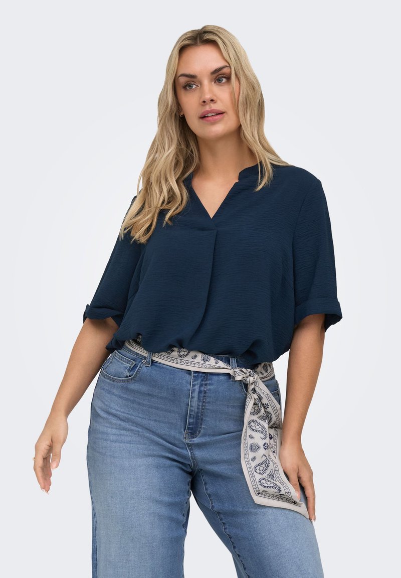 Woman wearing a dark blue short-sleeve blouse tucked into light blue jeans with a patterned scarf belt, standing against a plain background.