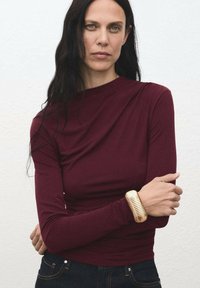 Burgundska bluza s dolgimi rokavi z drapirano zasnovo, kombinirana s temnim denimom. Na zapestju je teksturiran zlati   bracelet, ki ustvarja vizualni kontrast.