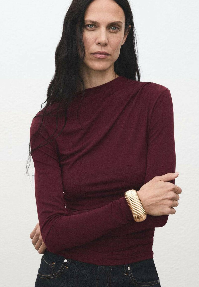 Burgundska bluza s dolgimi rokavi z drapirano zasnovo, kombinirana s temnim denimom. Na zapestju je teksturiran zlati   bracelet, ki ustvarja vizualni kontrast.
