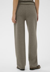 Osoba oblečená v voľných sivých nohaviciach a hnedých topánkach, stojaca proti jednofarebnému bielému pozadiu, z pohľadu zozadu.