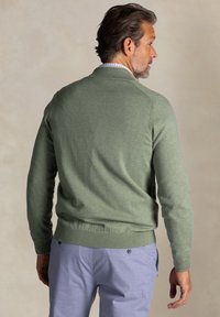 Groene gebreide cardigan met een ribgebreide zoom, gedragen over een overhemd met kraag, in combinatie met lichtblauwe broek. Beschikt over een eenvoudige, gladde textuur.