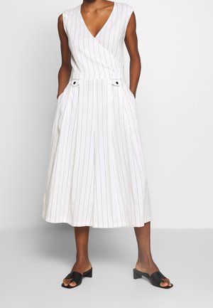 Weißes, gestreiftes Kleid mit V-Ausschnitt und ärmellosem Design, mit tailliertem Schnitt und seitlichen Taschen. Schwarze, blockabsatzige Pantoletten.