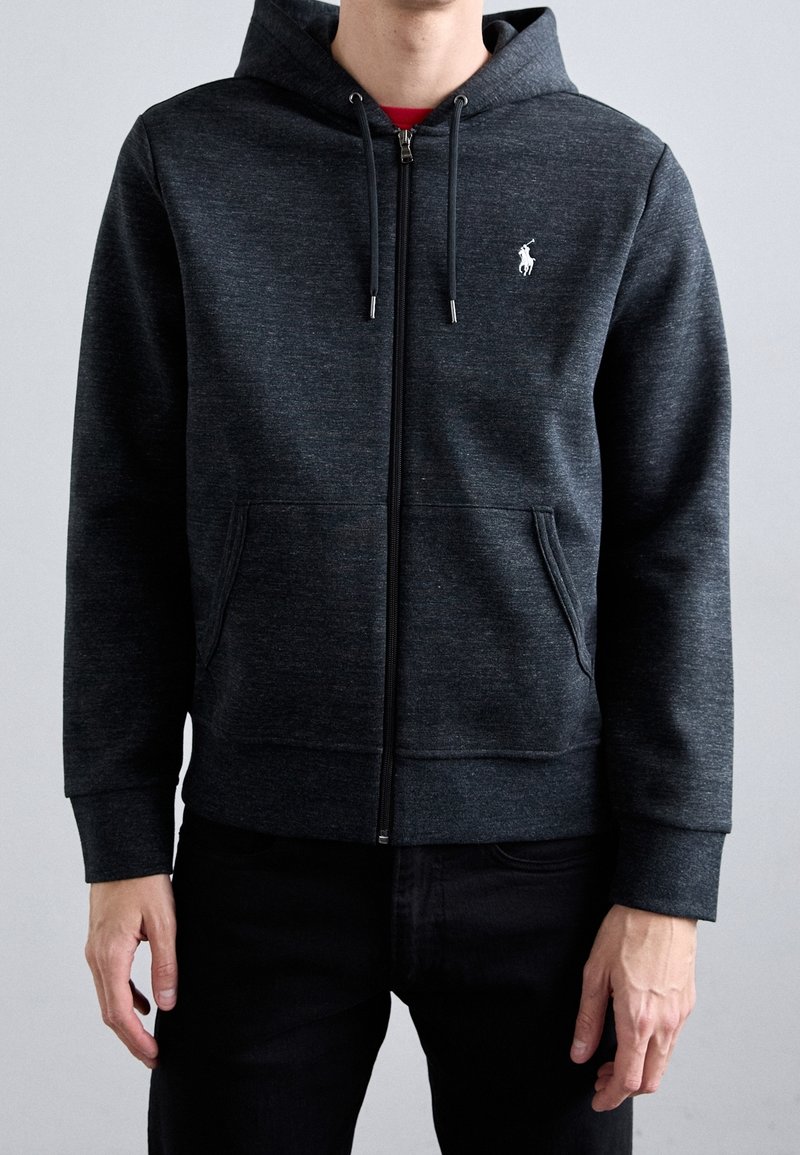 Homme portant un sweat à capuche zippé gris foncé avec poches avant et petit logo blanc sur le côté gauche de la poitrine, assorti à un pantalon noir.