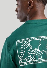 Green cotton t-shirt featuring a white graphic on the back with playful figures and text "POP SHOP TOKYO" and "POP SHOP NYC" in a rectangular border.
