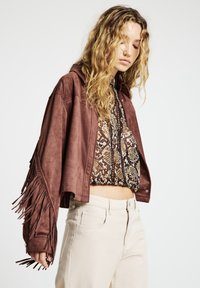 Braune Wildlederjacke in kurzer Form mit Fransenverzierung an den Ärmeln, getragen über einem gemusterten Oberteil. Hellfarbene, eng anliegende Hosen vervollständigen den Look.