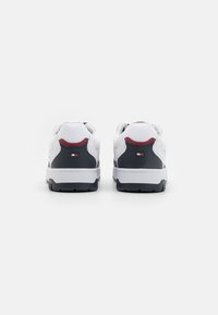 Sneakers bianchi in pelle con accenti grigi, caratterizzati da un dettaglio rosso sul tallone e una toppa con logo. Suola in gomma spessa con motivo antiscivolo.