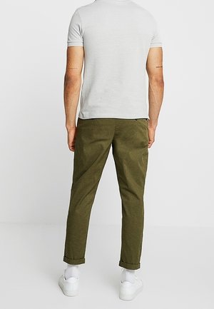 Chemise grise à manches courtes avec un col rond, associée à un pantalon chino vert olive. Les pantalons ont des ourlets retroussés et sont fabriqués dans un tissu doux et texturé. Baskets blanches.