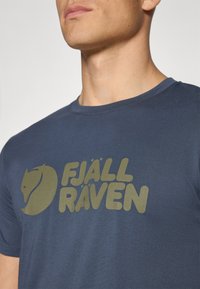 Tmavomodré bavlnené tričko s veľkým svetlozeleným logom "FJÄLL RÄVEN" a siluetou líšky, s okrúhlym výstrihom a krátkymi rukávmi.