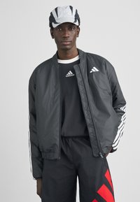 Zwarte bomberjack met witte Adidas-logo's en zijstrepen, gecombineerd met een zwart shirt en een grijze en zwarte pet. Gestructureerde stof, strak design.