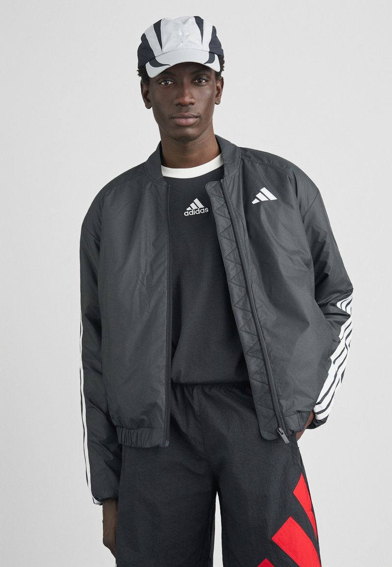 Schwarze Bomberjacke mit weißen Adidas-Logos und seitlichen Streifen, kombiniert mit einem schwarzen Shirt und einer grau-schwarzen Kappe. Strukturiertes Gewebe, elegantes Design.