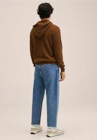 Bruine geribde hoodie met een capuchon, gecombineerd met lichtblauwe baggy jeans en beige sneakers, gezien van achteren tegen een neutrale achtergrond.