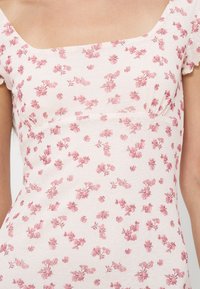 Personne portant une robe rose pâle à encolure carrée et manches courtes, avec un motif floral rose sur l’ensemble de la robe.
