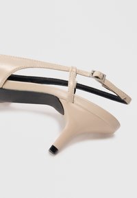 Beige high-heeled shoe with a glossy finish, open-toe design, and a slender ankle strap featuring a small metallic buckle. Black sole accent.