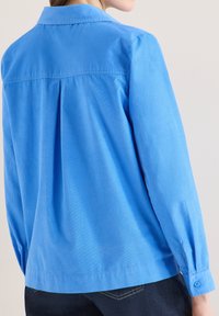 Femme portant une chemise bleu clair à manches longues avec un pli au dos, associée à un jean bleu foncé, vue de dos devant un mur uni.