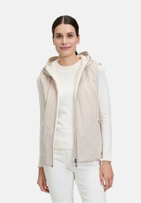 Veste légère sans manches en beige avec une capuche, équipée d'une fermeture éclair à double sens et d'une doublure intérieure douce. Tissu lisse avec des détails de design minimaux.