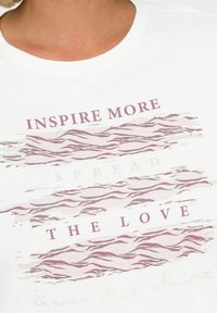 Bílý tričko s textem: "INSPIRUJTE VÍCE," "ROZŠIŘTE," a "LÁSKU" v různých písmech; obsahuje abstraktní vlnité vzory v tlumené růžové a zelené.