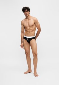 Homme athlétique debout pieds nus portant un slip noir avec une ceinture blanche sur un fond blanc uni.
