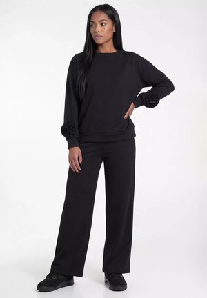 Kobieta stoi z ręką na biodrze, ubrana w czarną bluzę z marszczonymi mankietami, szerokie czarne spodnie i czarne trampki na białym tle.