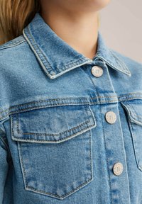 Gaiši zila džinsu jaka ar klasisku apkakli, diviem krūšu maciņiem, vara akcentiem un kontrastējošiem dūrieniem uz strukturētas auduma.