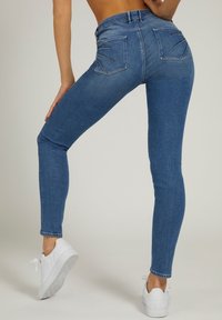 Jeans de denim azul ajustados con una cintura alta, que presentan dos bolsillos traseros y un patrón texturizado. Combinados con zapatillas blancas.