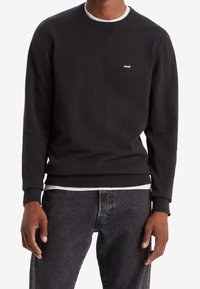 Svart tröja i bomullsblandning med rundad halsringning, ribbade muddar och en liten vit Levi's-logotyp på bröstet. Buren över grå jeans.