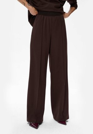 Wijdvallende broek in donkerbruin stof, met een elastische tailleband en een soepele textuur. Geaccentueerd met puntige, burgundy hakken.