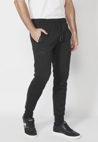 Černé jogger kalhoty vyrobené z měkké tkaniny, s bočními kapsami na zip, s elastickým pasem na šňůrku a se zužujícími se nohavicemi. Nosené s černými teniskami.