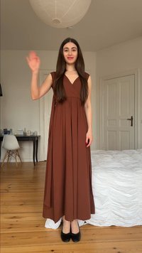 Femme aux longs cheveux bruns portant une robe marron et des chaussures noires faisant signe dans une chambre avec un sol en bois et des meubles blancs.