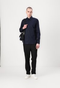 Tengerész fűzőgombos ing, hosszú ujjú, slim fit, fekete nadrággal és fehér sportcipővel párosítva. A modellen egy kis fekete táska van.