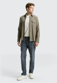 Licht olijfgroene knop-up overhemd met twee voorzakken, gedragen over een wit T-shirt, gecombineerd met blauwe spijkerbroeken en witte sneakers.