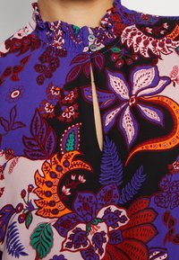 Lila blus med blommönster som har en volangkrage, knappst ängning och livfulla flerfärgade mönster med gröna och röda accenter.