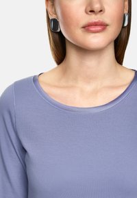 Light purple long-sleeve top with a rounded neckline and smooth texture, paired with large silver and black square earrings.