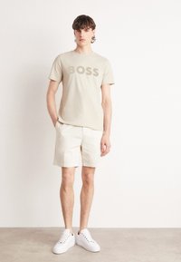 Beige színű póló "BOSS" felirattal, kontrasztos árnyalatban, krém színű sorttal és fehér sportcipővel párosítva, egy modellel, aki egy egyszínű háttér előtt áll.