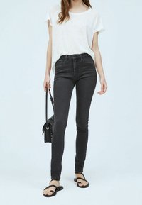 Jeans negros de cintura alta con un ajuste entallado, combinados con una camiseta blanca holgada y sandalias negras. Se incluye un pequeño bolso negro de hombro.