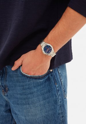 Person iført et sølvfarvet metalur med en blå urskive, hånd i lomme, iført en mørk langærmet skjorte og blå jeans.