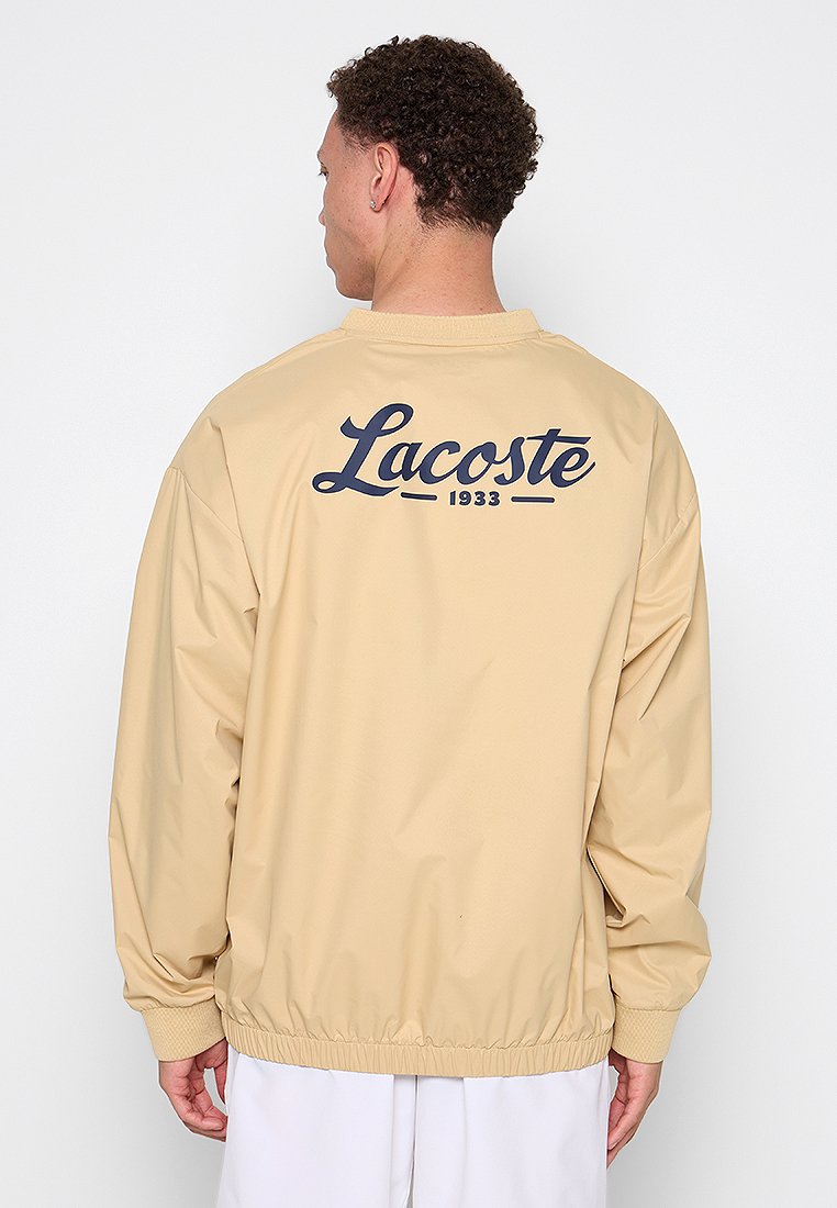 Veste beige légère avec des poignets et un ourlet côtelés. Présente le logo "Lacoste 1933" en bleu foncé au dos. Texture lisse, coupe décontractée.