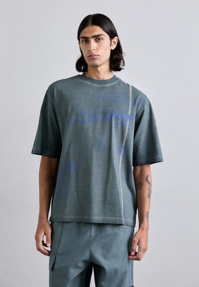 T-shirt vert à manches courtes avec un col rond. Il présente un design de texte bleu fané et une couture diagonale en accent. Fabriqué en tissu doux et texturé.