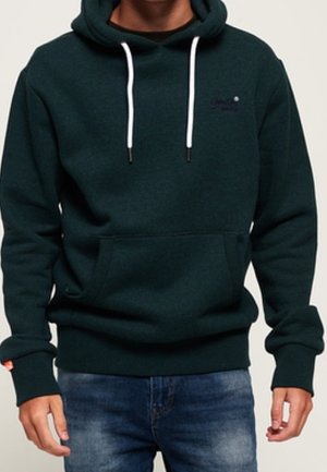Mann trägt dunkelgrünes Kapuzensweatshirt mit weißen Kordeln und Fronttasche, kombiniert mit blauen Jeans.
