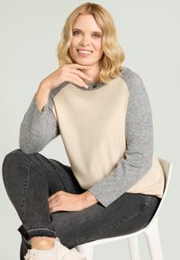 Bluza w kolorze beżowym i szarym z blokowym wzorem, miękką teksturą, szarymi rękawami oraz ozdobami przy dekolcie, noszona z ciemnymi dżinsami.