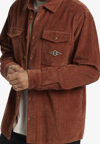 Rust corduroy shirt with vertical ribbing, two chest pockets, front buttons, and embroidered diamond patch featuring wave design.