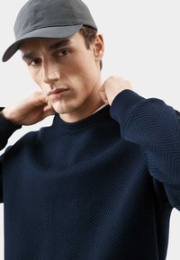 Sudadera texturizada azul marino con un patrón de chevron, cuello redondo y puños acanalados. Combinada con una gorra de béisbol de pana gris.