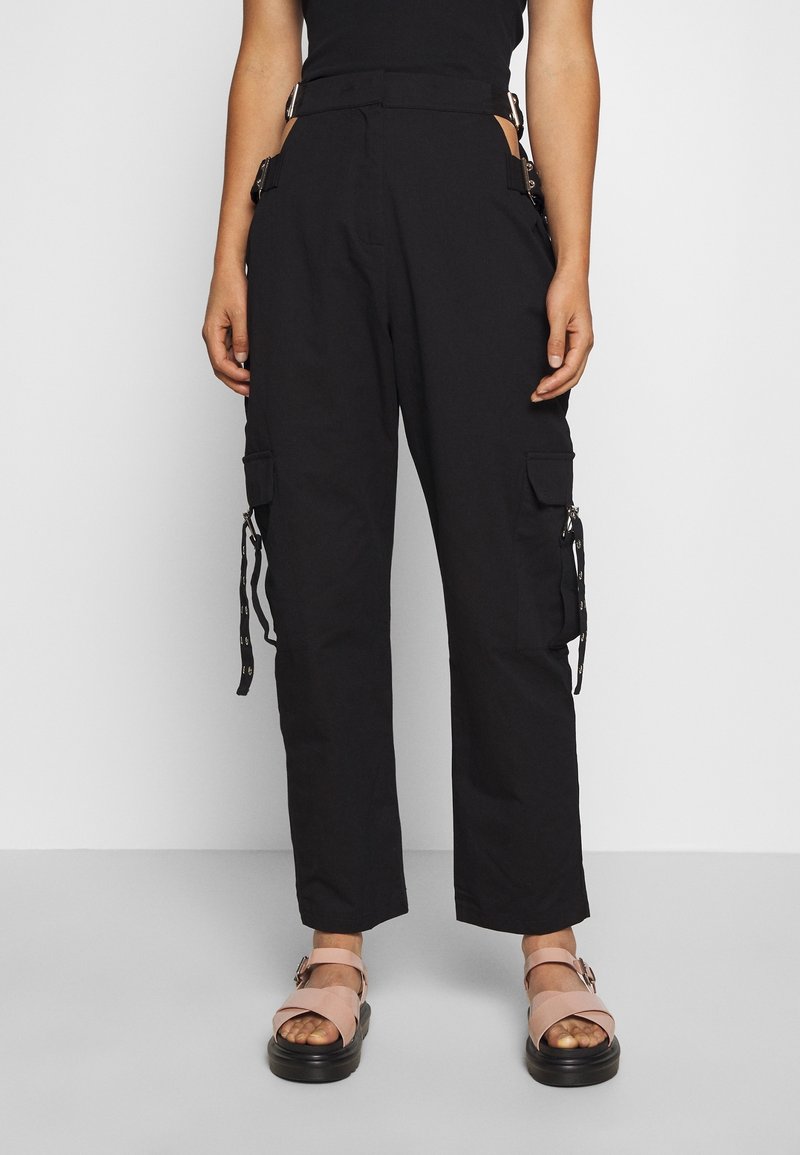 Femme portant un pantalon cargo noir avec des poches latérales et des sandales à plateforme roses, debout devant un fond blanc uni.