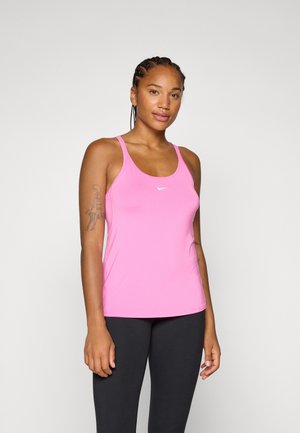 Kvinde med fletninger, iført en pink Nike tanktop og sorte leggings, står mod en neutral baggrund og smiler selvsikkert.