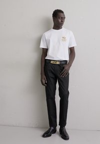 Camiseta blanca con un logo de oso de peluche, pantalones negros y un cinturón dorado "MOSCHINO". El calzado son zapatos negros, pulidos. Diseño simple y ajustado.