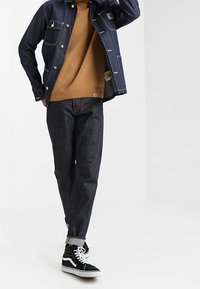 Kurtka dżinsowa z białymi szwami, brązowy sweter, ciemne dżinsy oraz czarne sneakersy za kostkę z białymi akcentami. Model stoi z uniesioną jedną ręką.