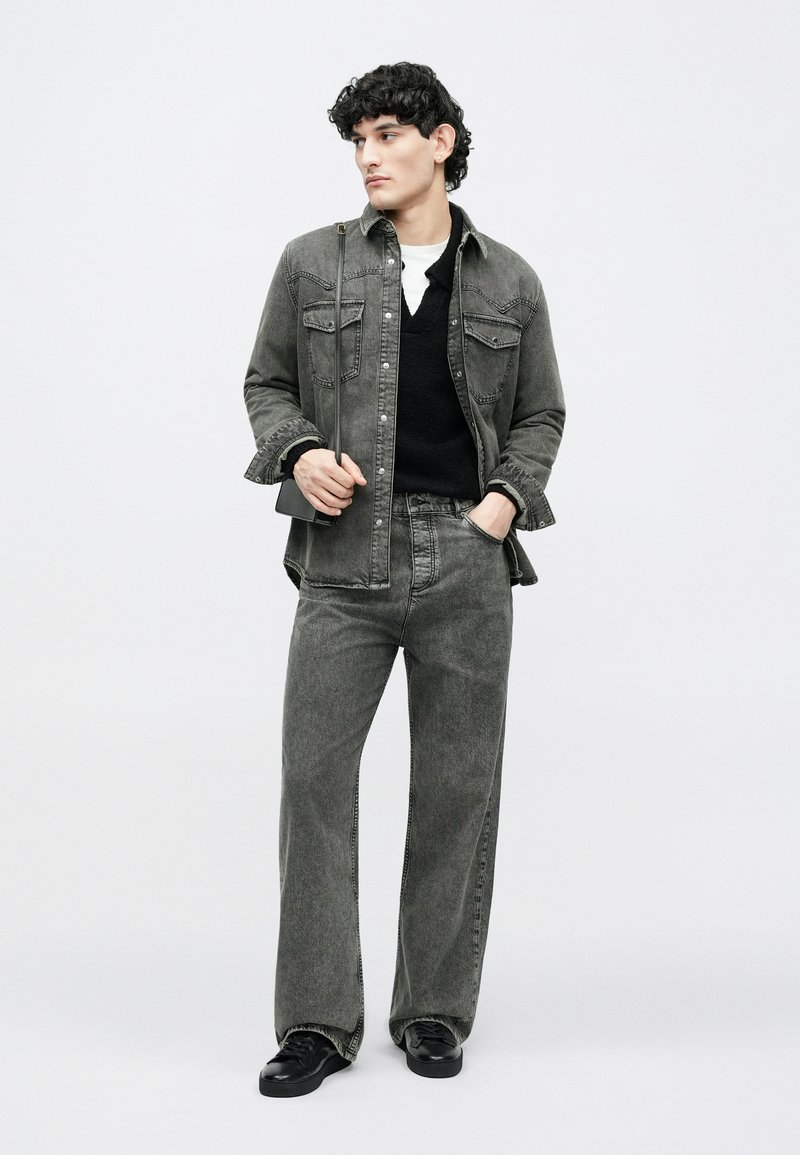 Young man with curly hair wearing faded black denim shirt and jeans, black sweater, white undershirt, black sneakers, holding small crossbody bag.